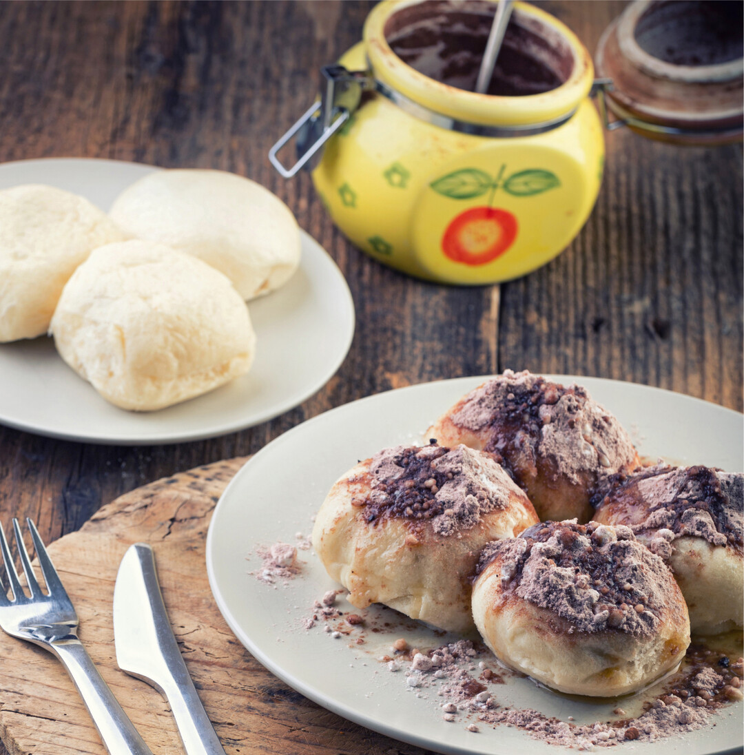 Damit Dampfnudeln die leckere, karamellartige Kruste bekommen, braucht es Aufmerksamkeit (Rezept folgende Seite). Milch, Butter und Zucker im Bratentopf erhitzen, die Hälfte der Nudeln dicht hineinsetzen, mit Deckel schließen, bei mittlerer Hitze garen. Immer wieder beobachten! Das Brodeln im Topf soll hörbar sein, Hitze anpassen. Nach 15 bis 20 Minuten lässt das Geräusch nach, dann knackt es und duftet nach Karamell. Nun darf der Deckel erstmals (!) abgenommen werden. Die zweite Hälfte backen Anfängerinnen vorsichtshalber im Ofen. Dampfnudeln schmecken auch mit Marmelade und Kakao (Bild).