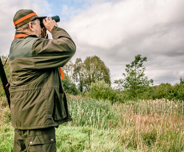 Ein Jger steht auf einer Wiese und sieht durch sein Fernglas.