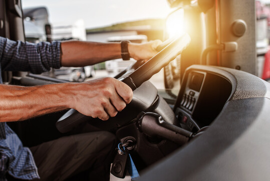 Fahrer eines Lkws mit beiden Hnden am Lenkrad und Sonnenreflexion im Auenspiegel.