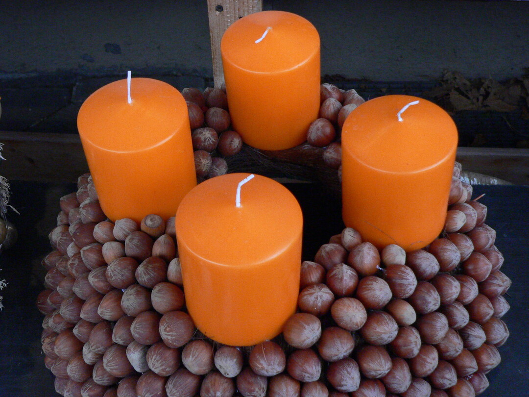 Mit Nüssen und Kerzen lässt sich ein einzigartiger Adventskranz zaubern. Die orangefarbenen Stumpen-kerzen in RAL-Qualität harmonieren dabei perfekt mit den Brauntönen der Nüsse.