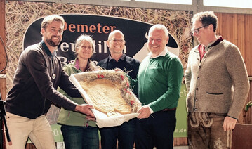 V.l.n.r.: Christoph Reiber (Geschftsfhrer Demeter Baden-Wrttemberg), Ute Haimerl (Geschftsfhrerin Demeter Bayern) und Alexander Gerber (Vorstand Demeter e.V.) berreichen Rolf Holzapfel und Andreas Aufmuth (beide Vorstnde bei den Demeter HeuMilch Bauern) auf der Jubilumsfeier in Ankenreute einen groen Keks in Form einer Kuh  gebacken von Demeter-Bckermeisterin Anne Dorer von der Eselsmhle.